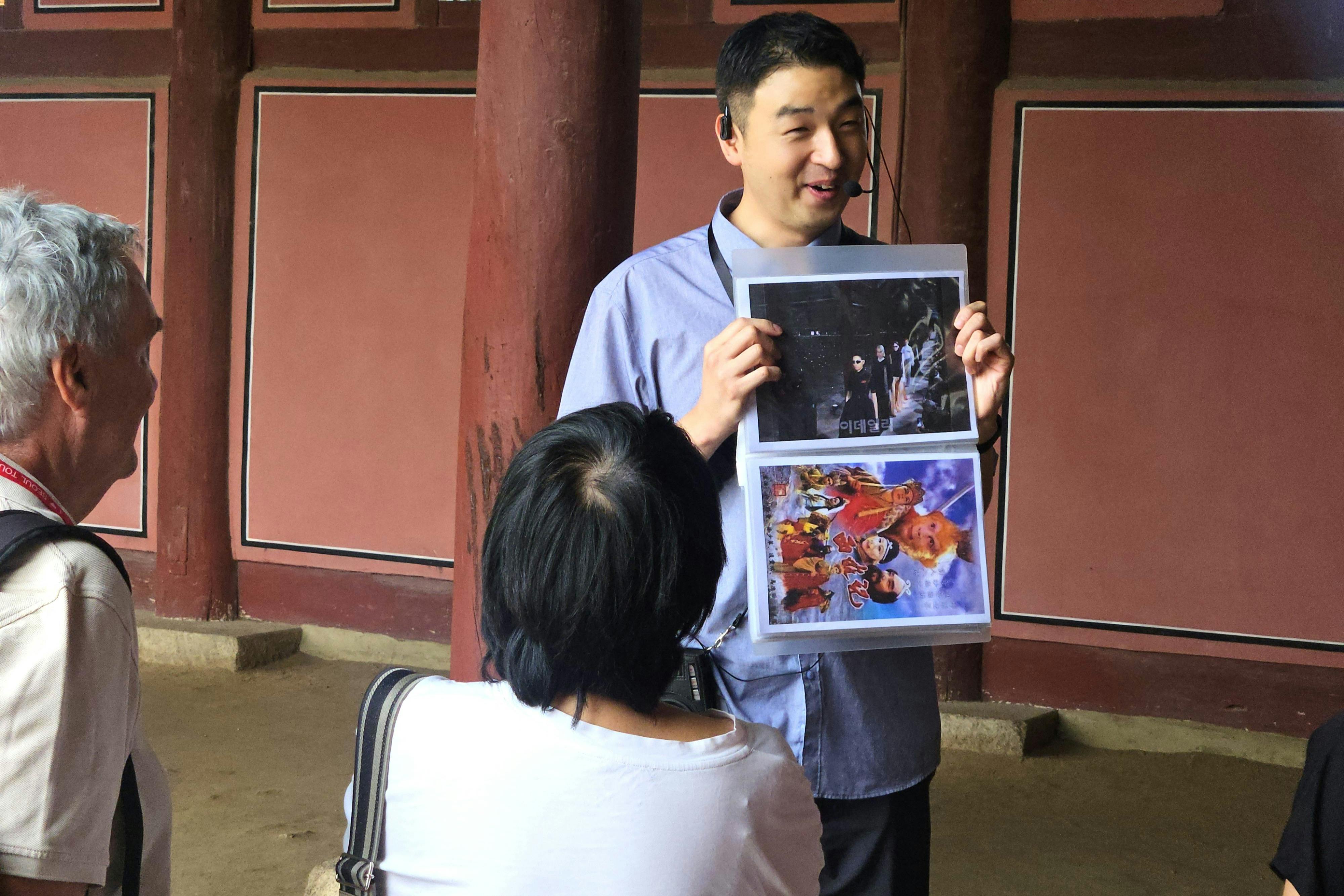 A person in a blue shirt is holding up a binder with images, addressing another seated person in an indoor setting.