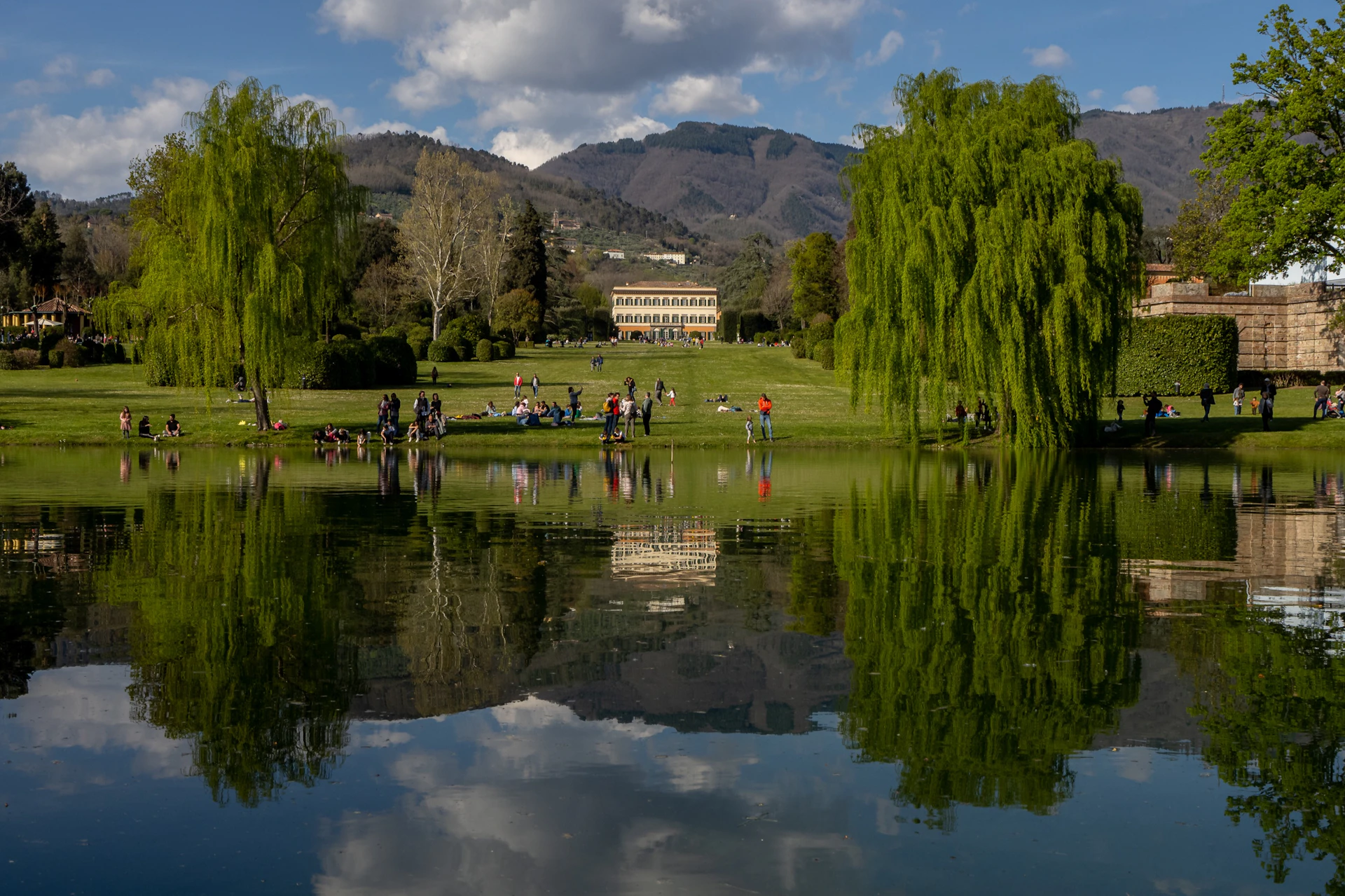 A serene park with a reflective pond, lush greenery, people relaxing, and a building in the background under a partly cloudy sky.
