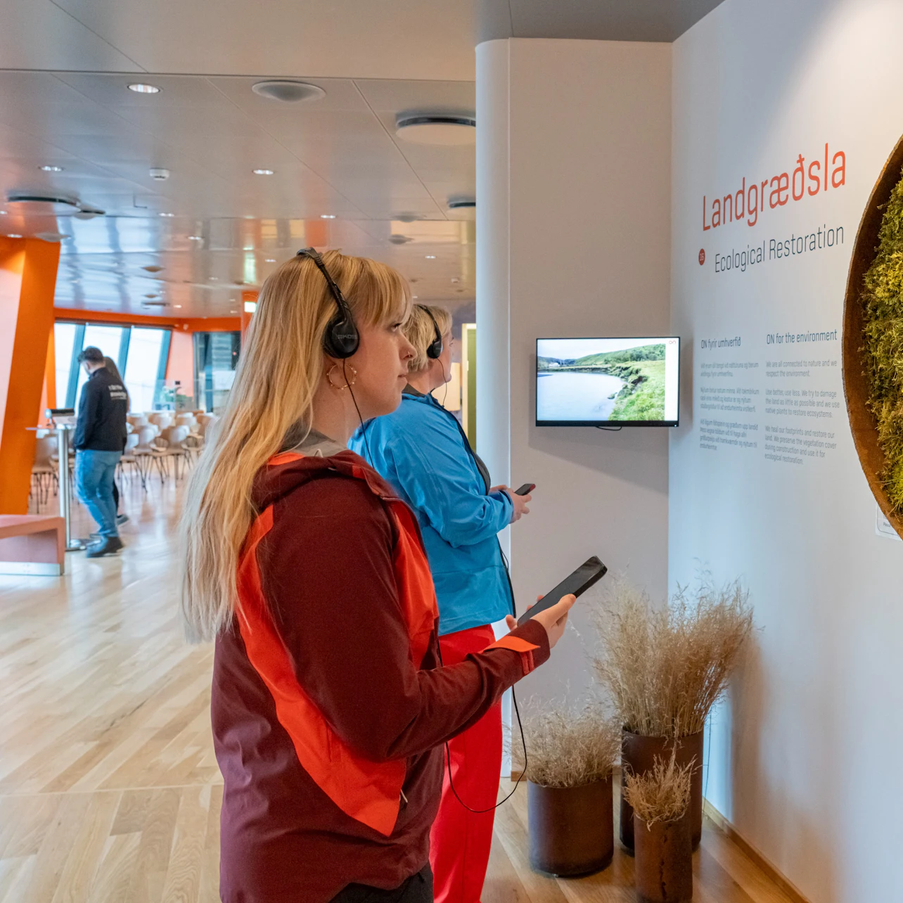 A woman in a red jacket wearing headphones interacts with an exhibit about ecological restoration indoors.