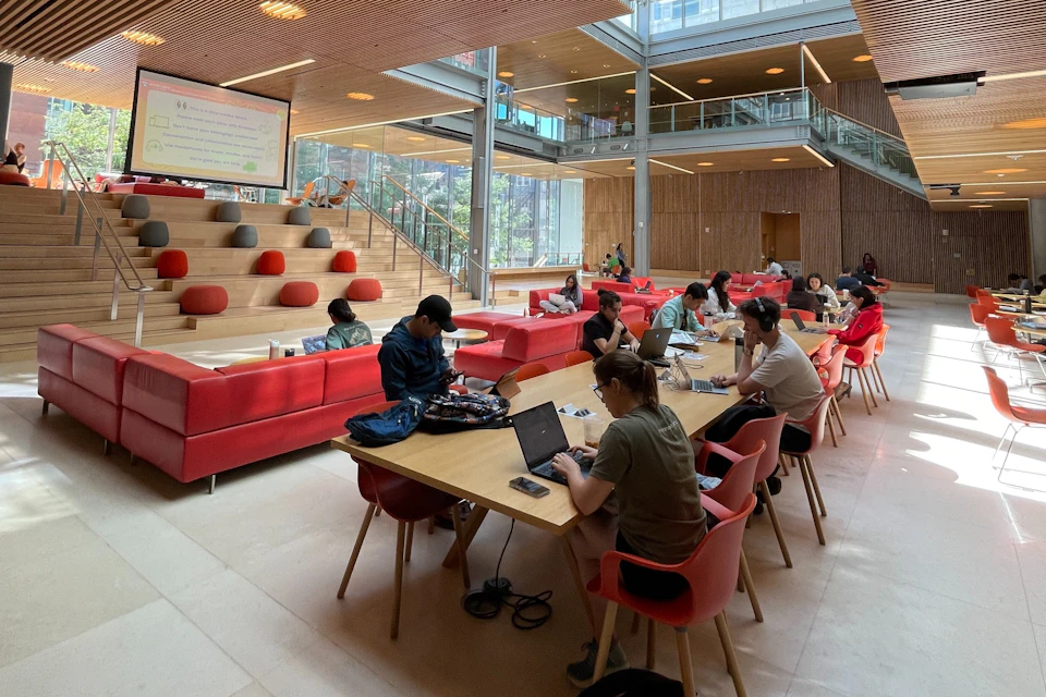 People working on laptops at long tables in a modern, open space with red couches, wooden accents, and large windows.