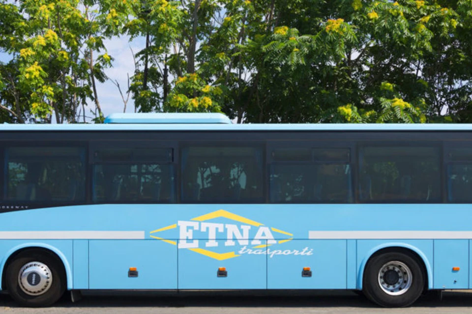 A blue bus with "ETNA trasporti" written on its side, parked in front of green trees with yellow flowers.