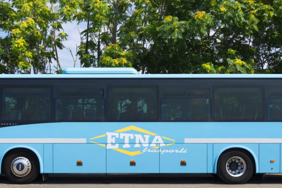 A blue bus with "ETNA trasporti" written on its side, parked in front of green trees with yellow flowers.