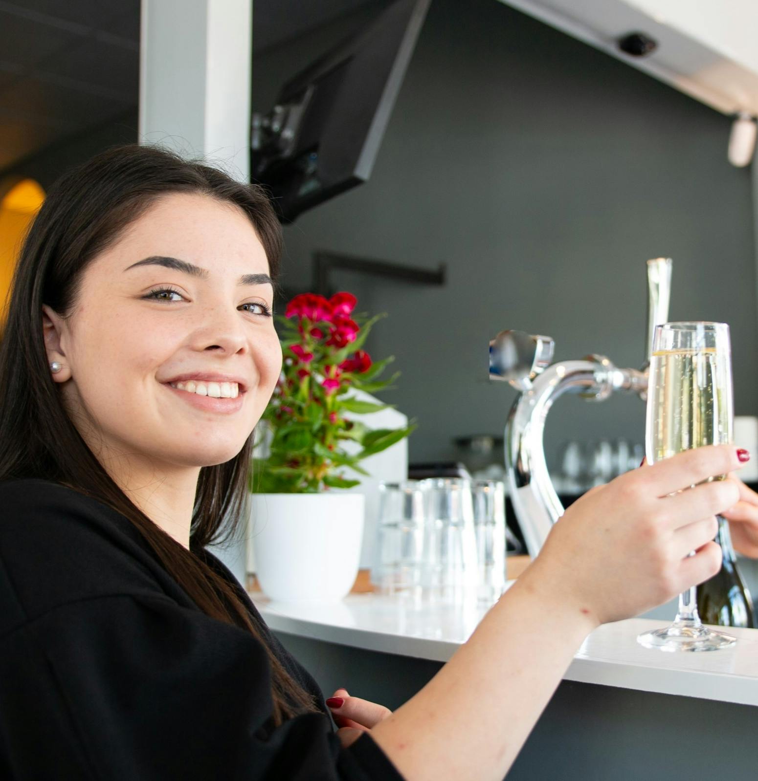 Μια γυναίκα χαμογελάει, κρατώντας ένα ποτήρι σαμπάνια σε ένα μπαρ με ένα φυτό και εργαλεία μπαρ στο παρασκήνιο.