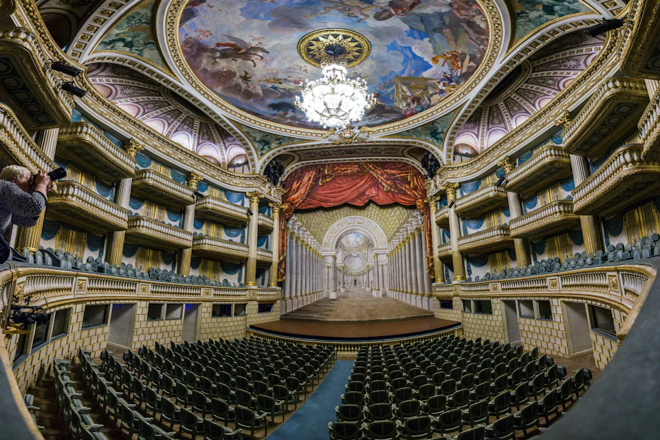 An ornate theater with a grand chandelier, painted ceiling, red curtain, stage, and rows of empty seats.