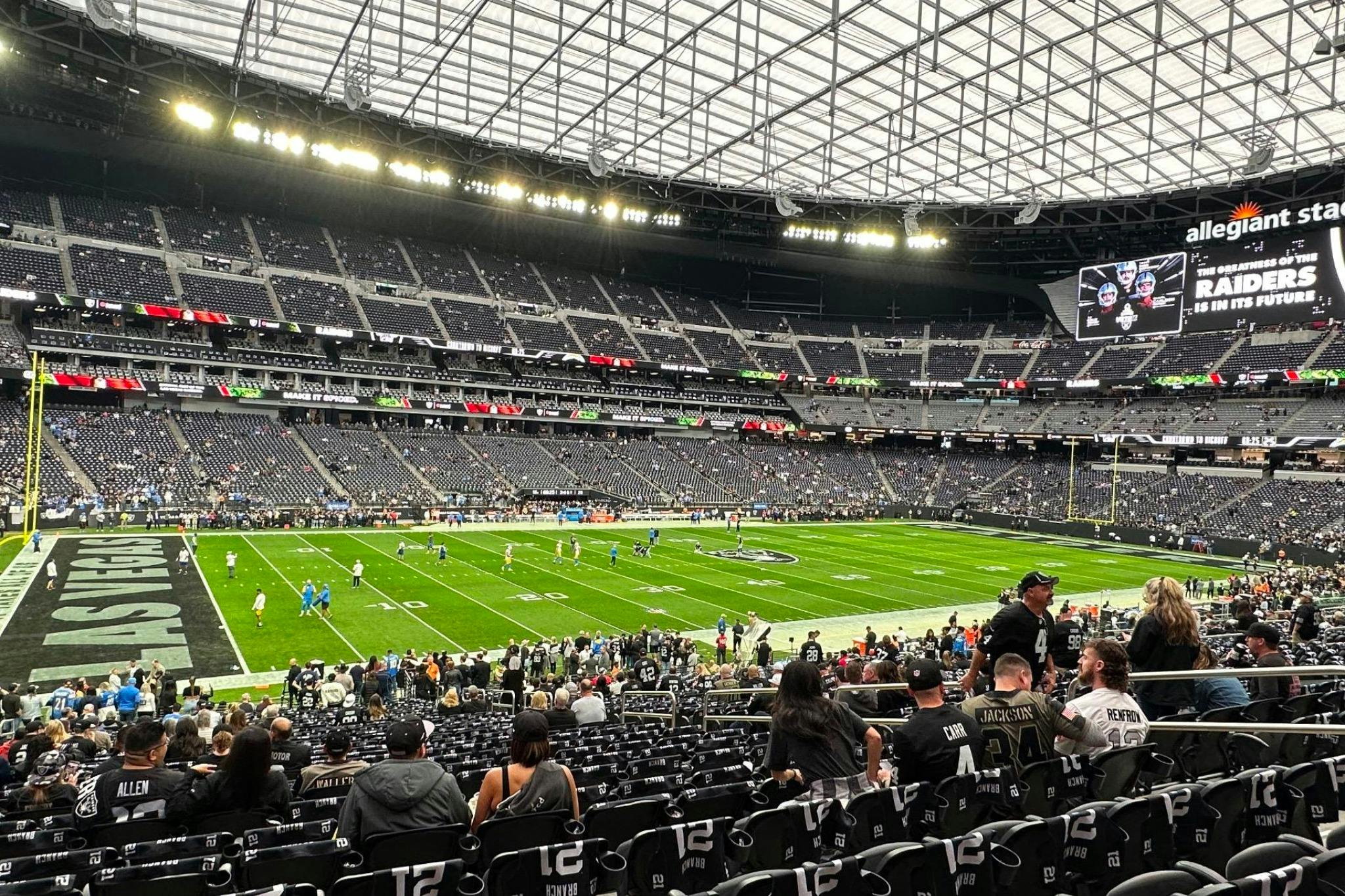 A football stadium with players warming up on the field and spectators in the stands, under a large roof structure.