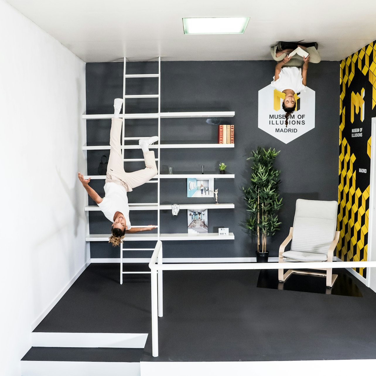 Två personer visas upp och ner i ett optiskt illusionsrum på Museum of Illusions Madrid, med möbler och hyllor på väggarna.