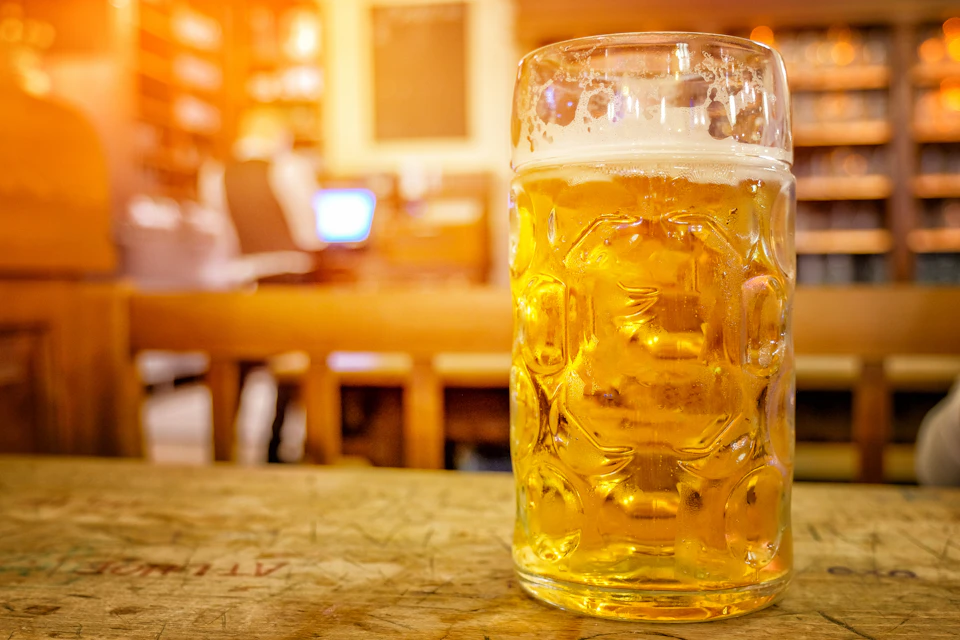 A large glass of beer on a wooden table with a blurred background of a person sitting and working on a laptop in a cozy setting.