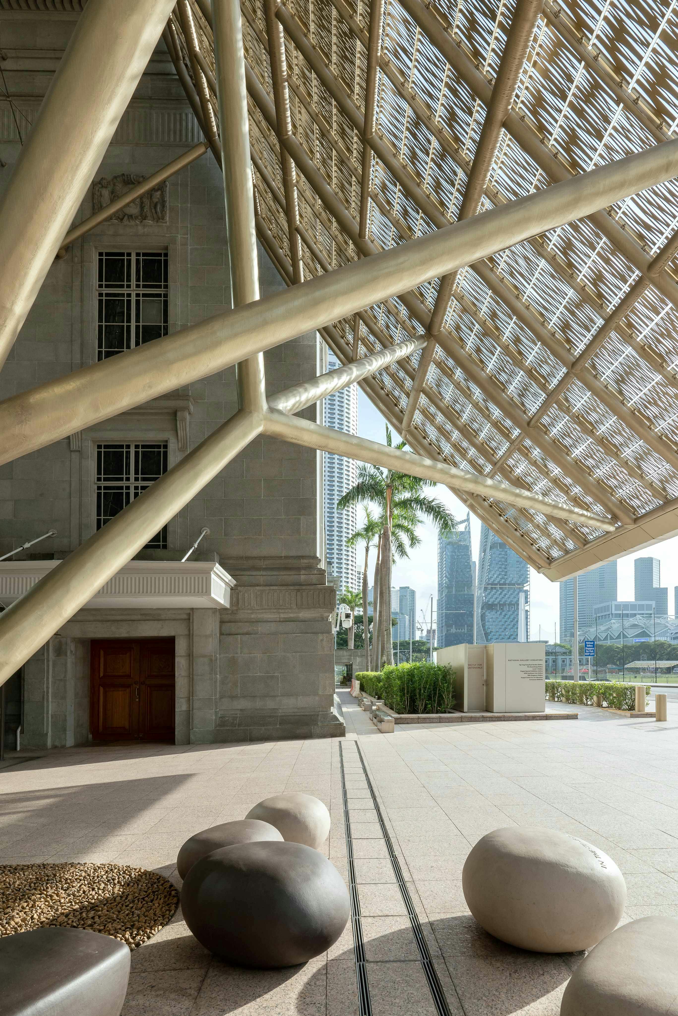Urban plaza with modern structures, large geometric roof, stone building, palm trees, and city skyscrapers in the background.