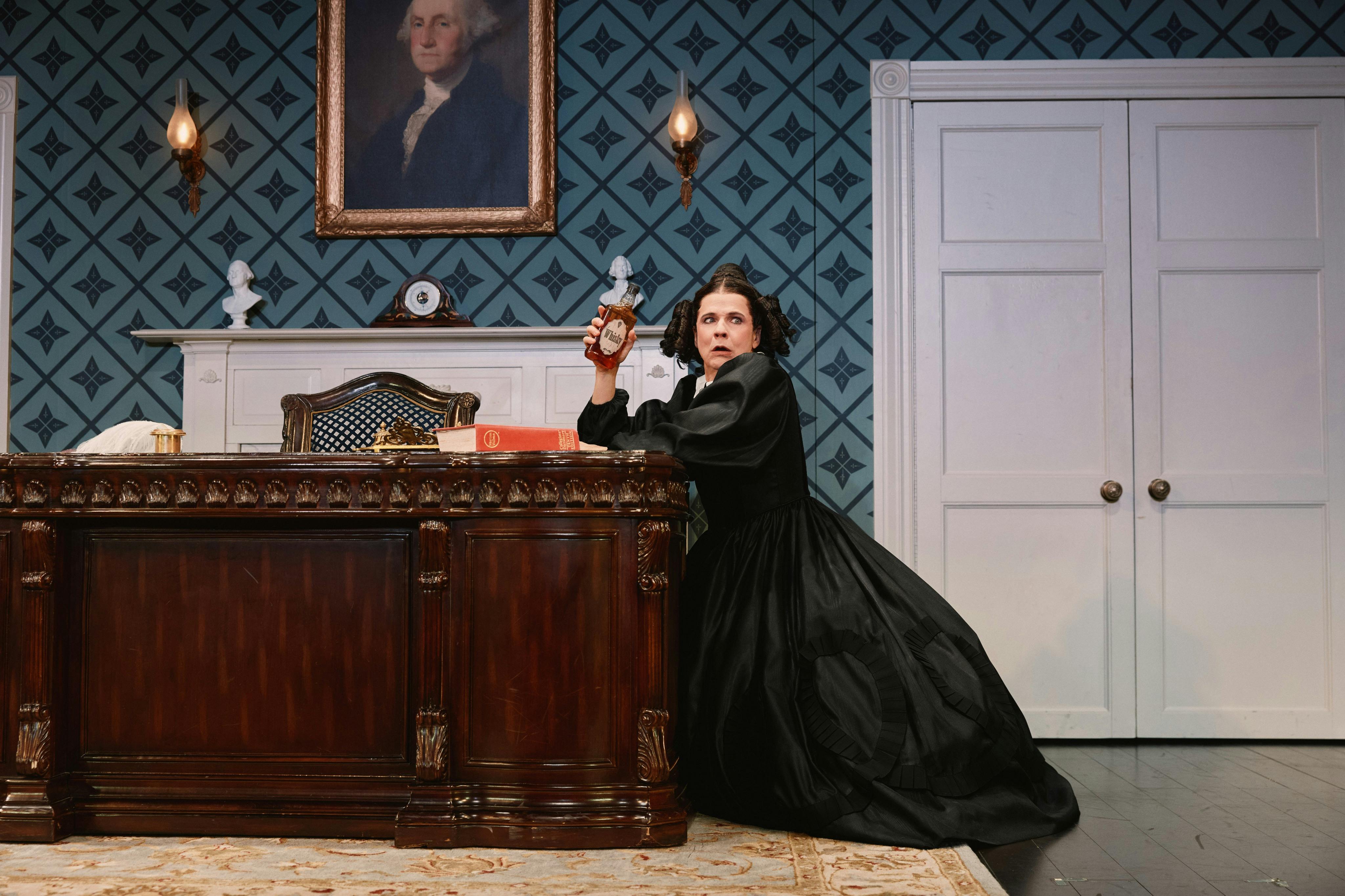 A person in a black dress crouches by a wooden desk holding an object, in a room with a patterned wall and a portrait above.