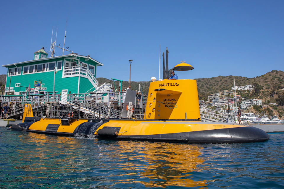 A yellow submarine named "Nautilus" is docked near a green building with stairs, with hills and buildings in the background.