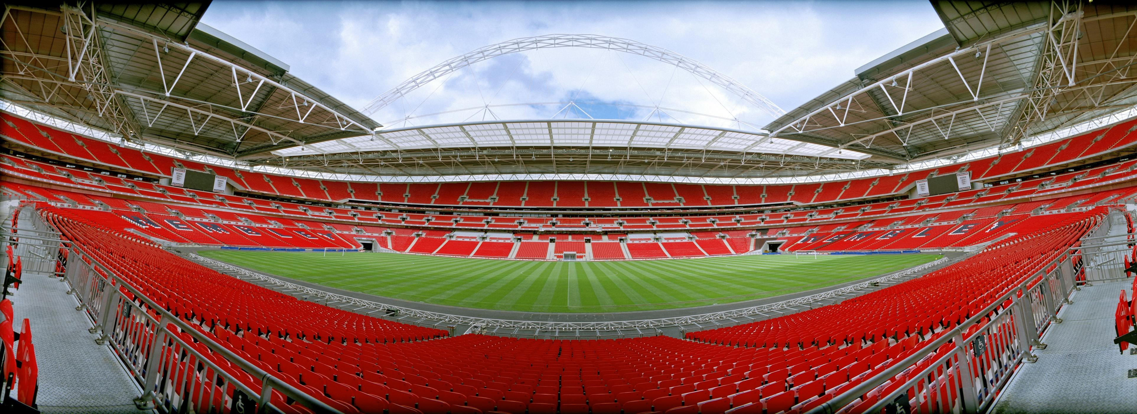 Panoramic view of an empty stadium with red seats surrounding a well-maintained green field, under a partially covered roof.