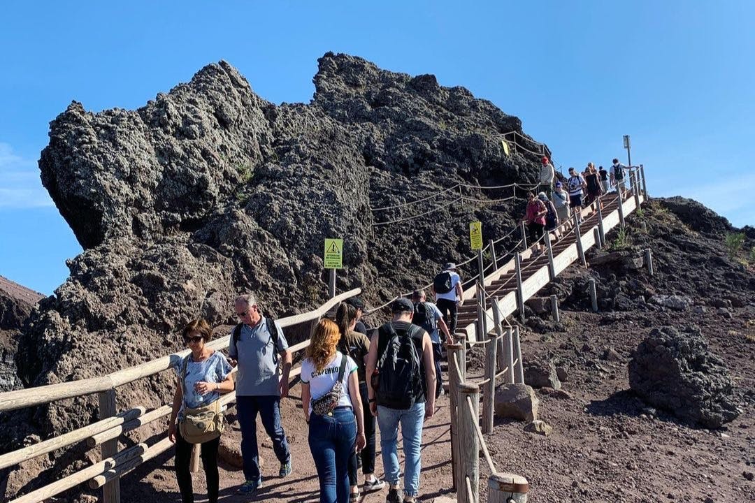Visitors climb a wooden pathway on a rocky mountain trail under a clear blue sky. Some signs are posted along the path.