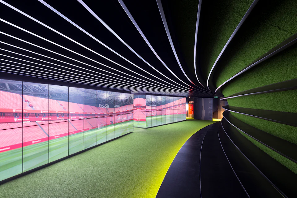 A modern hallway with curved walls, digital screens displaying red stadium images, and green flooring with overhead striped lighting.