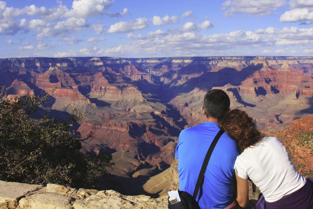 A couple sitting at the edge of the Grand Canyon, admiring the expansive view of the canyon under a partly cloudy sky.