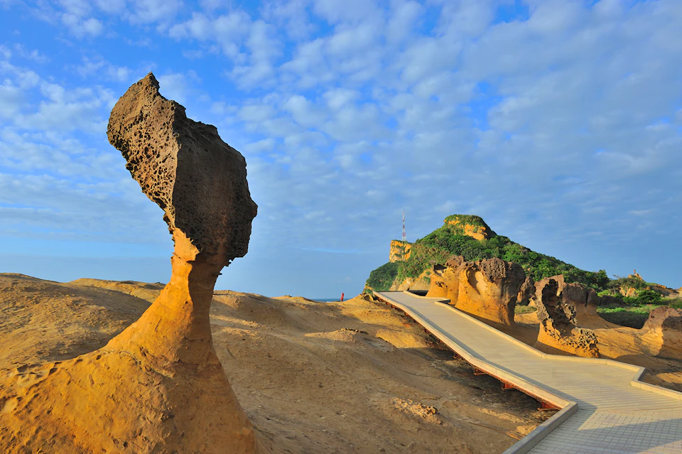 Rock formations with unique shapes, a wooden pathway, and a hill covered in greenery under a partly cloudy sky.