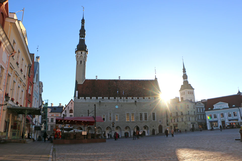 Town square with historic buildings, including a large structure with a tall spire, and a few people walking under a clear, sunny sky.