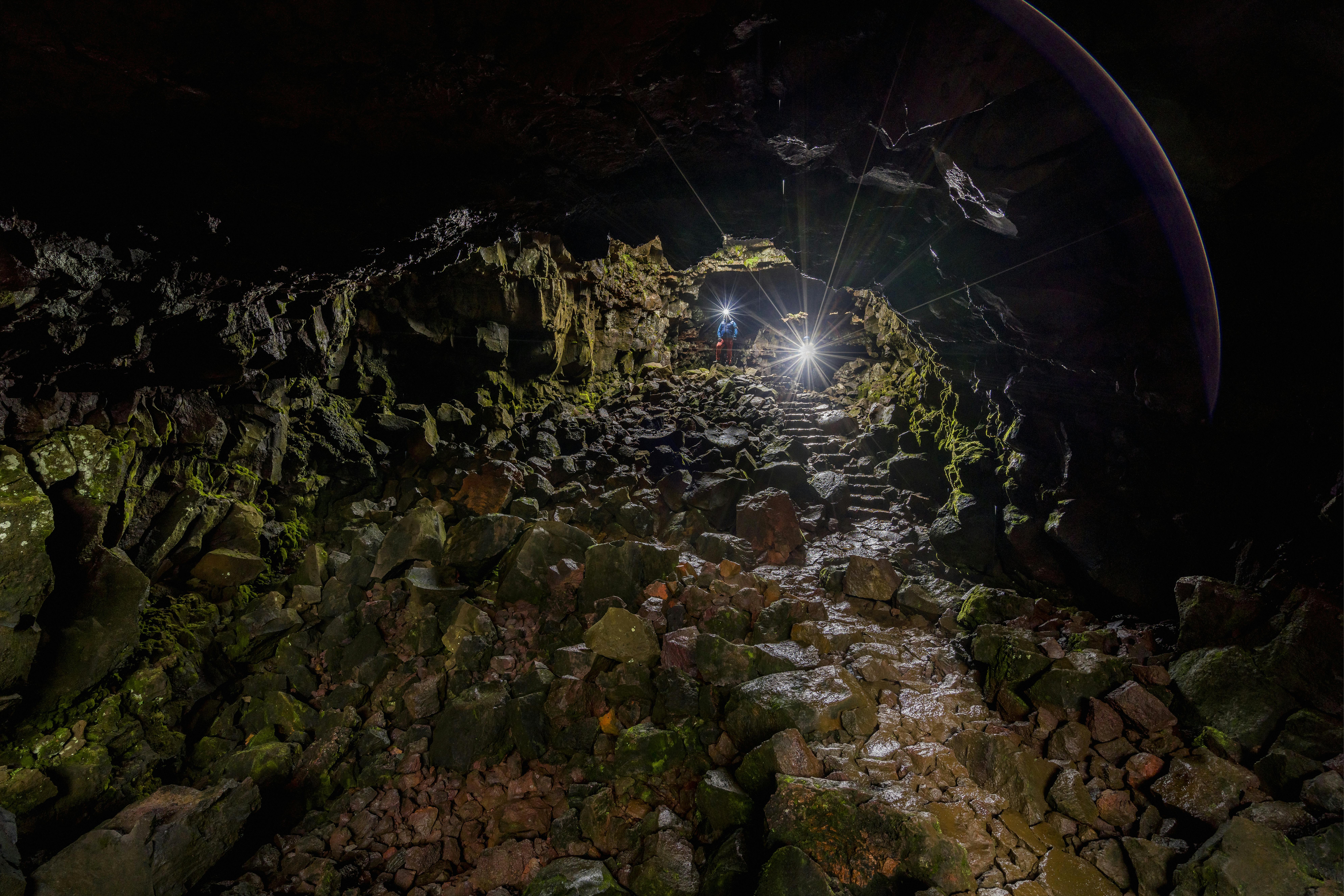 Person with headlamp standing in a rocky cave with light beaming out from an opening above.