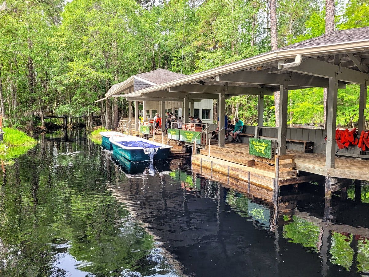 Et træbådshus med mennesker sidder på en rolig flod, omgivet af tætte grønne træer. Flere både ligger til kaj ved molen.