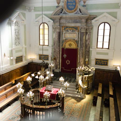An ornate synagogue interior with chandeliers, wooden benches, a red Torah ark curtain, and Hebrew inscriptions on the walls.