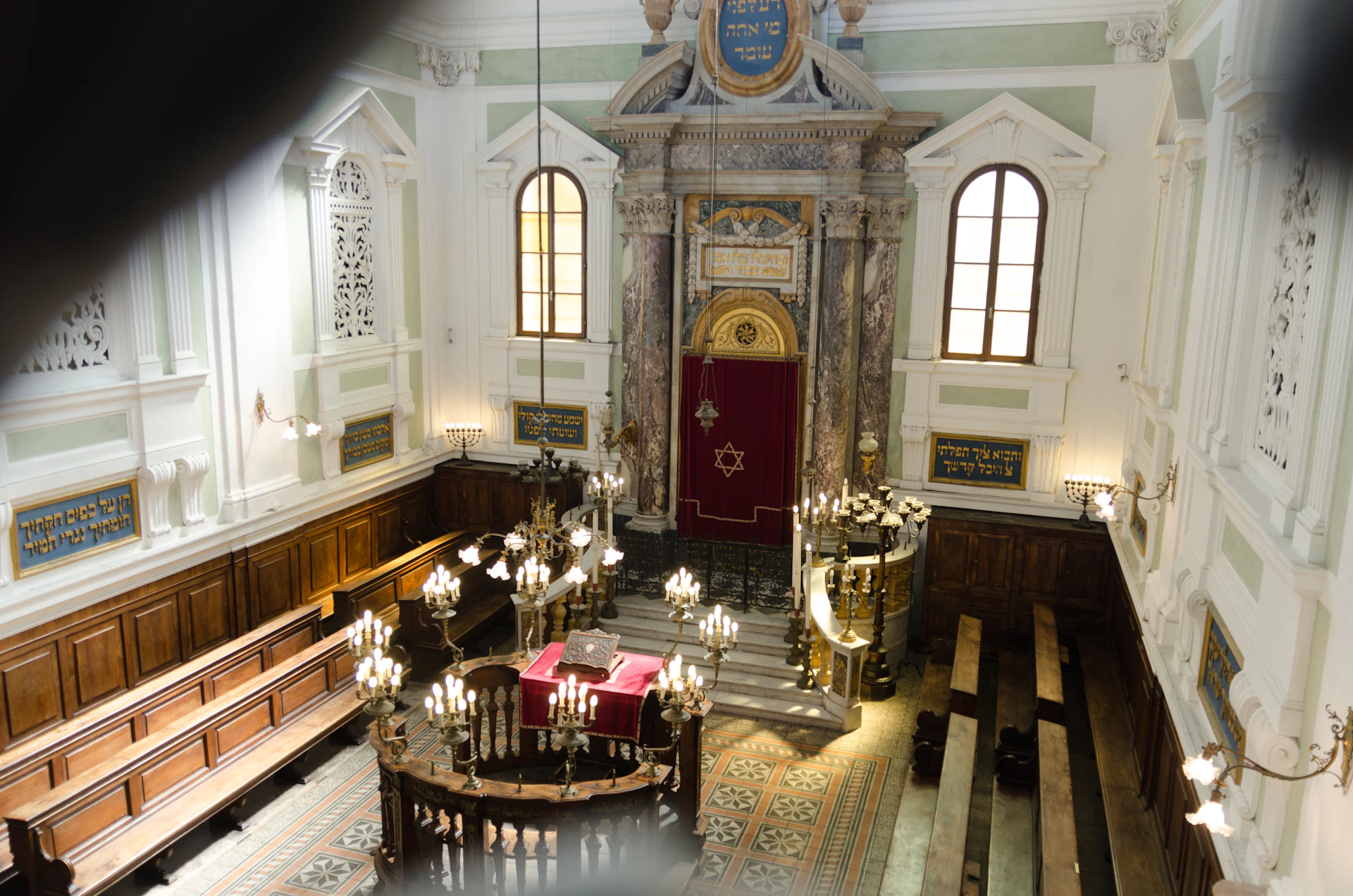 An ornate synagogue interior with chandeliers, wooden benches, a red Torah ark curtain, and Hebrew inscriptions on the walls.