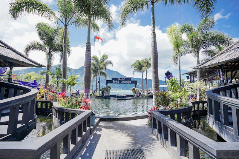 A picturesque resort entrance featuring a pathway leading to a pool surrounded by palm trees, with mountains in the background.