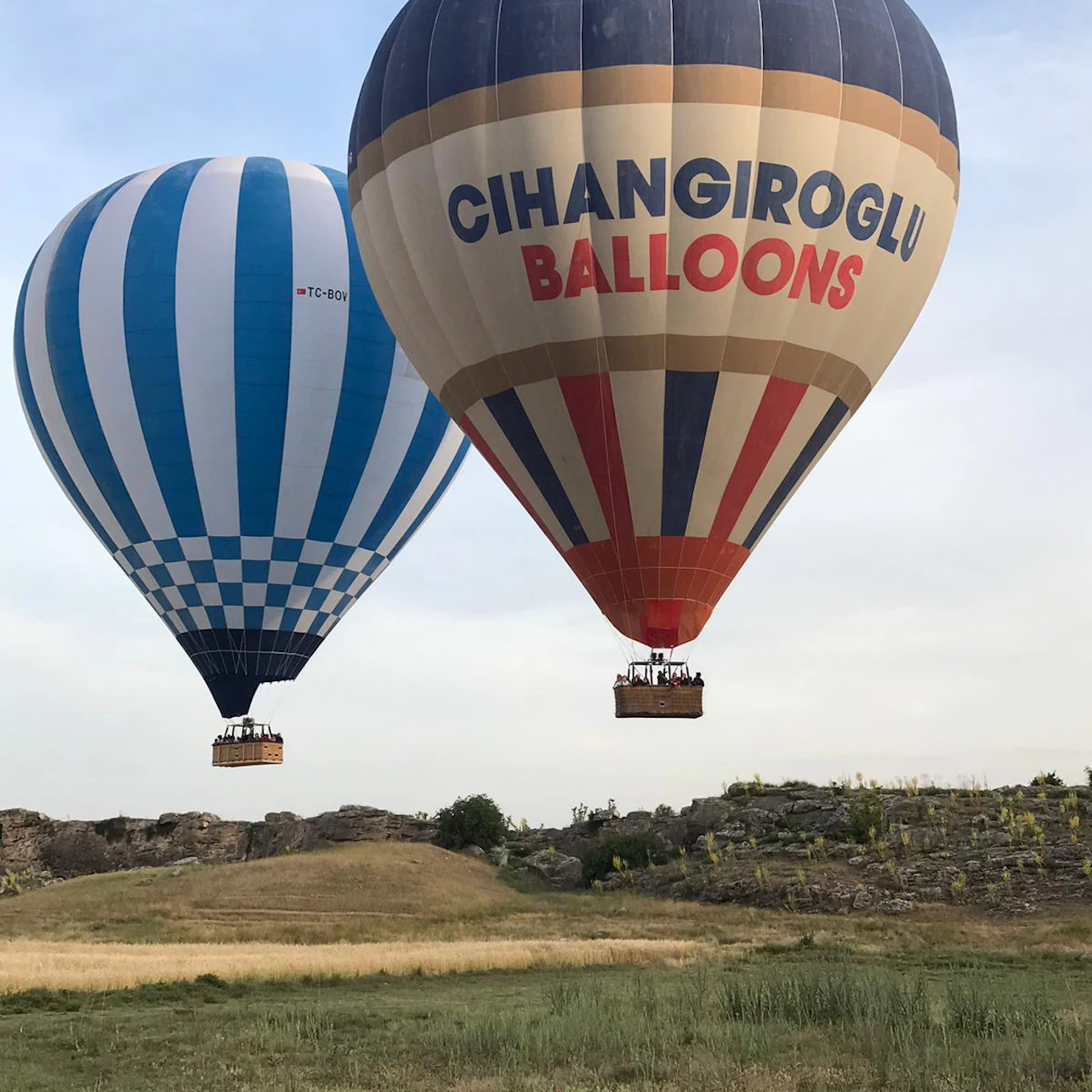 Two hot air balloons, one with blue stripes and the other marked "CIHANGIROGLU BALLOONS," soar above a grassy, rocky landscape.