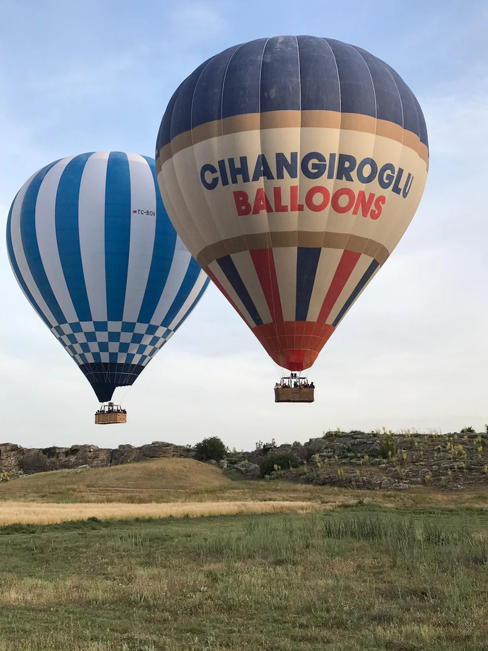 Two hot air balloons, one with blue stripes and the other marked "CIHANGIROGLU BALLOONS," soar above a grassy, rocky landscape.