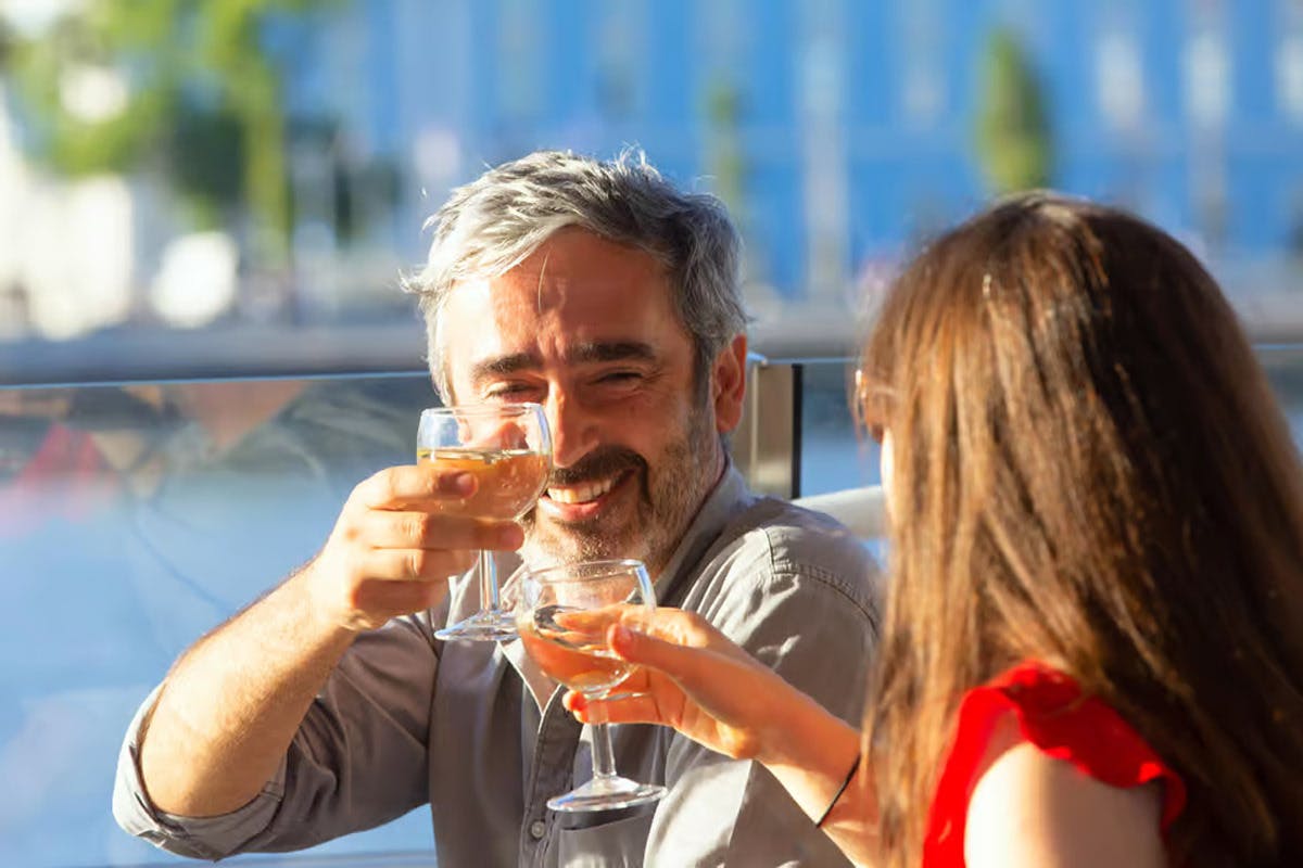 한 남자와 한 여자가 흐릿한 도시 배경의 야외에 앉아 미소를 지으며 와인 잔을 쥐고 있습니다.