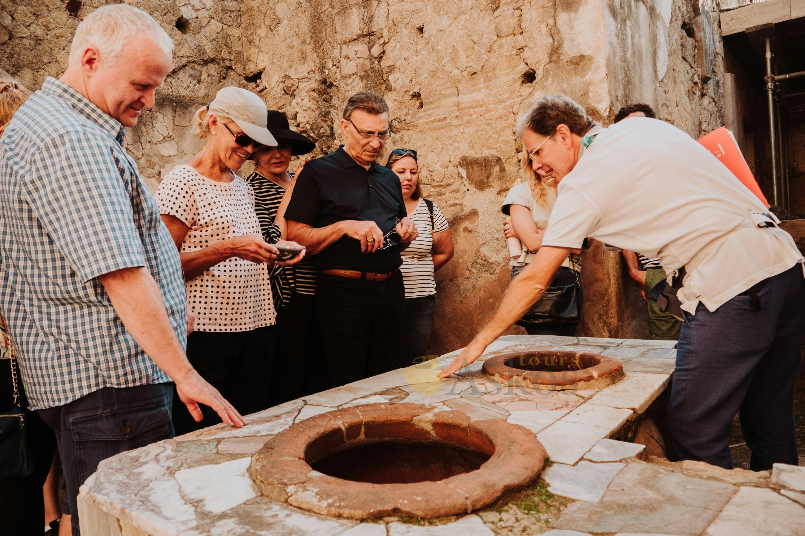 石のテーブルに円形の窪みがある歴史的な石造りの建物でツアーガイドの話を聞く人々のグループ。