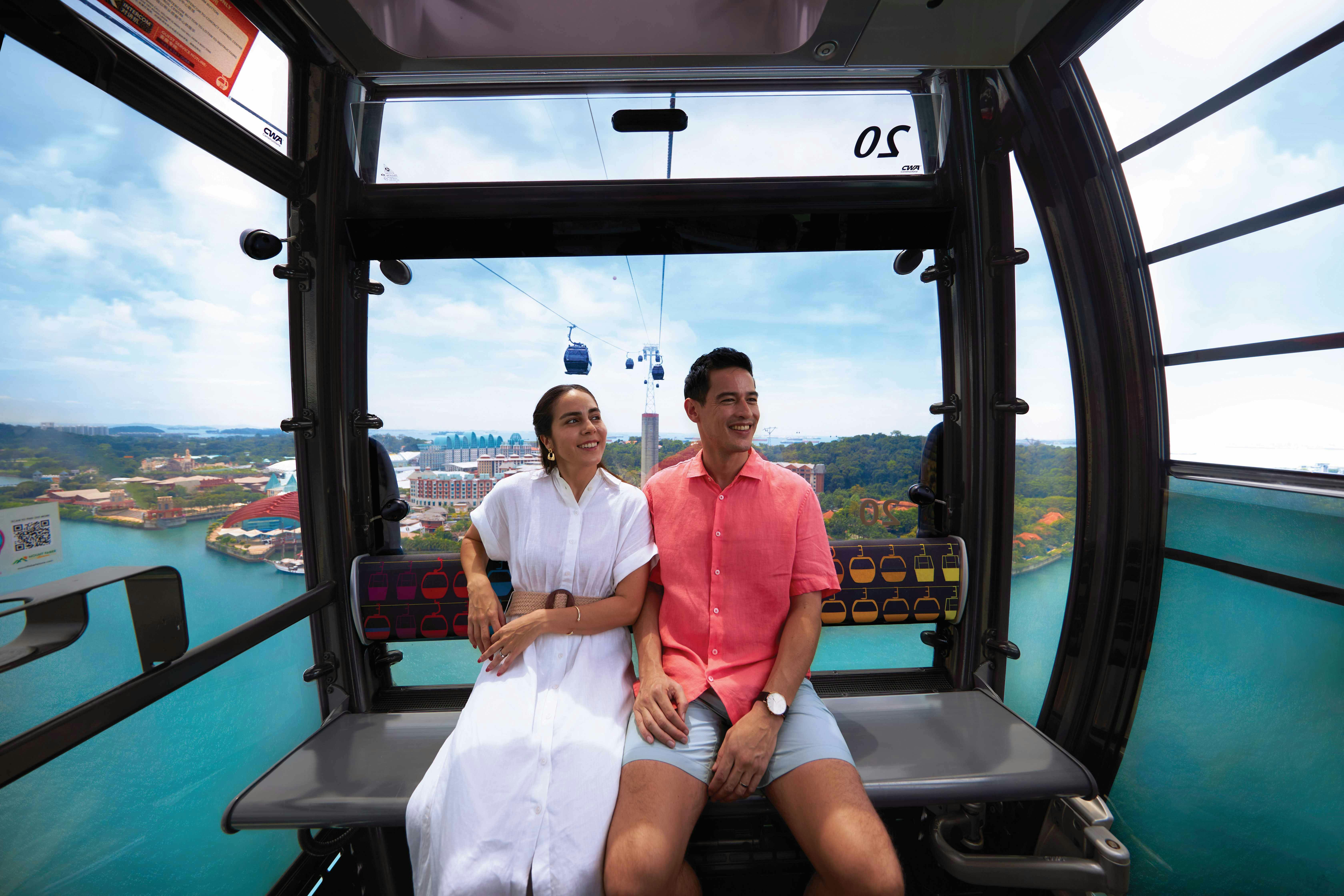 Ένα χαμογελαστό ζευγάρι κάθεται μέσα σε ένα τελεφερίκ με θέα τον ορίζοντα της πόλης και το καταπράσινο τοπίο με τα ήρεμα γαλάζια νερά από κάτω.
