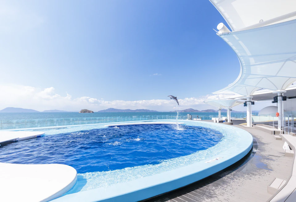 A dolphin jumps out of a circular pool by the ocean under a clear blue sky, with distant islands and modern architecture visible.