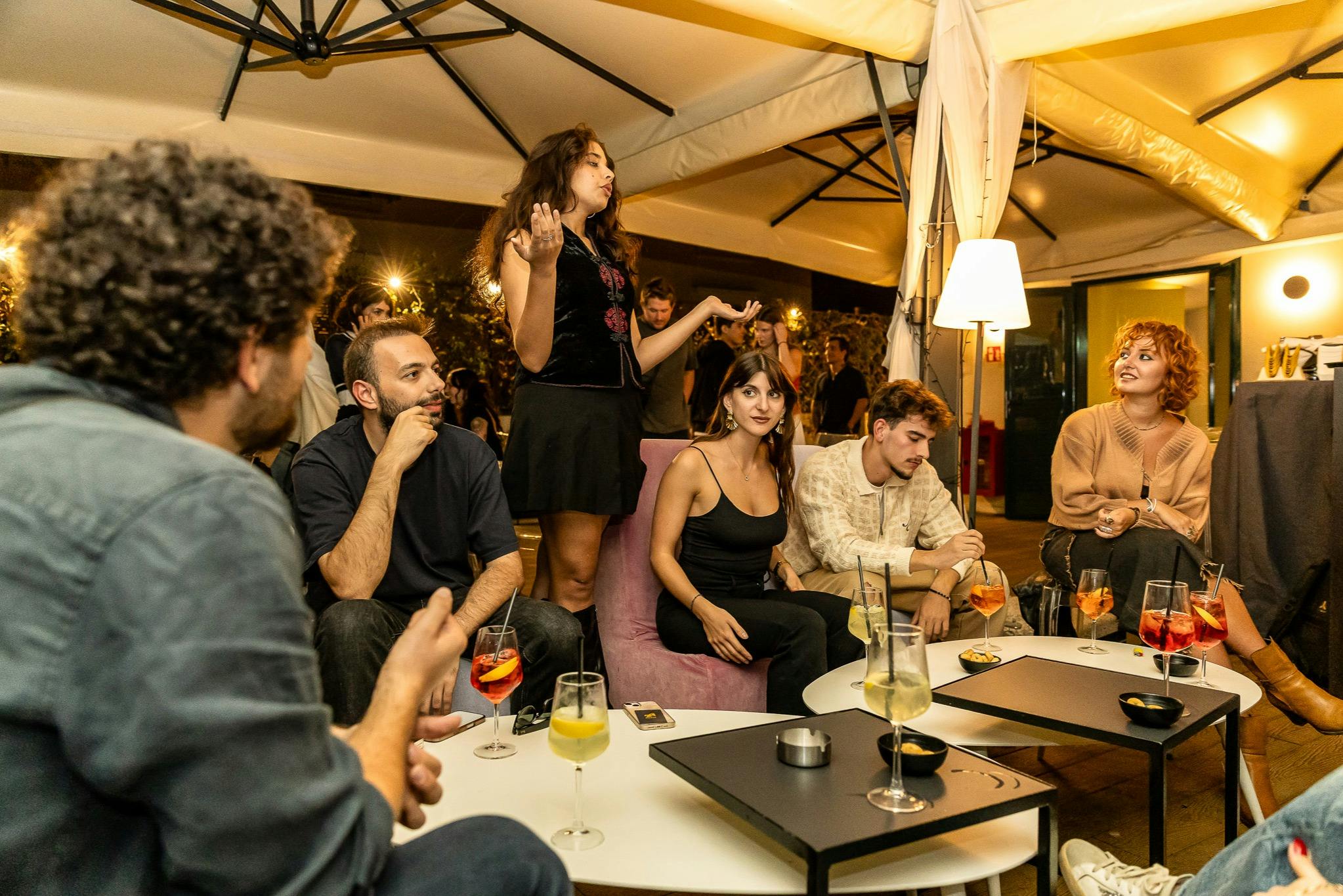 캐노피 아래에서 음료를 마시며 테이블에서 사람들과 어울리는 한 무리의 사람들, 한 사람이 서서 열정적으로 제스처를 취합니다.