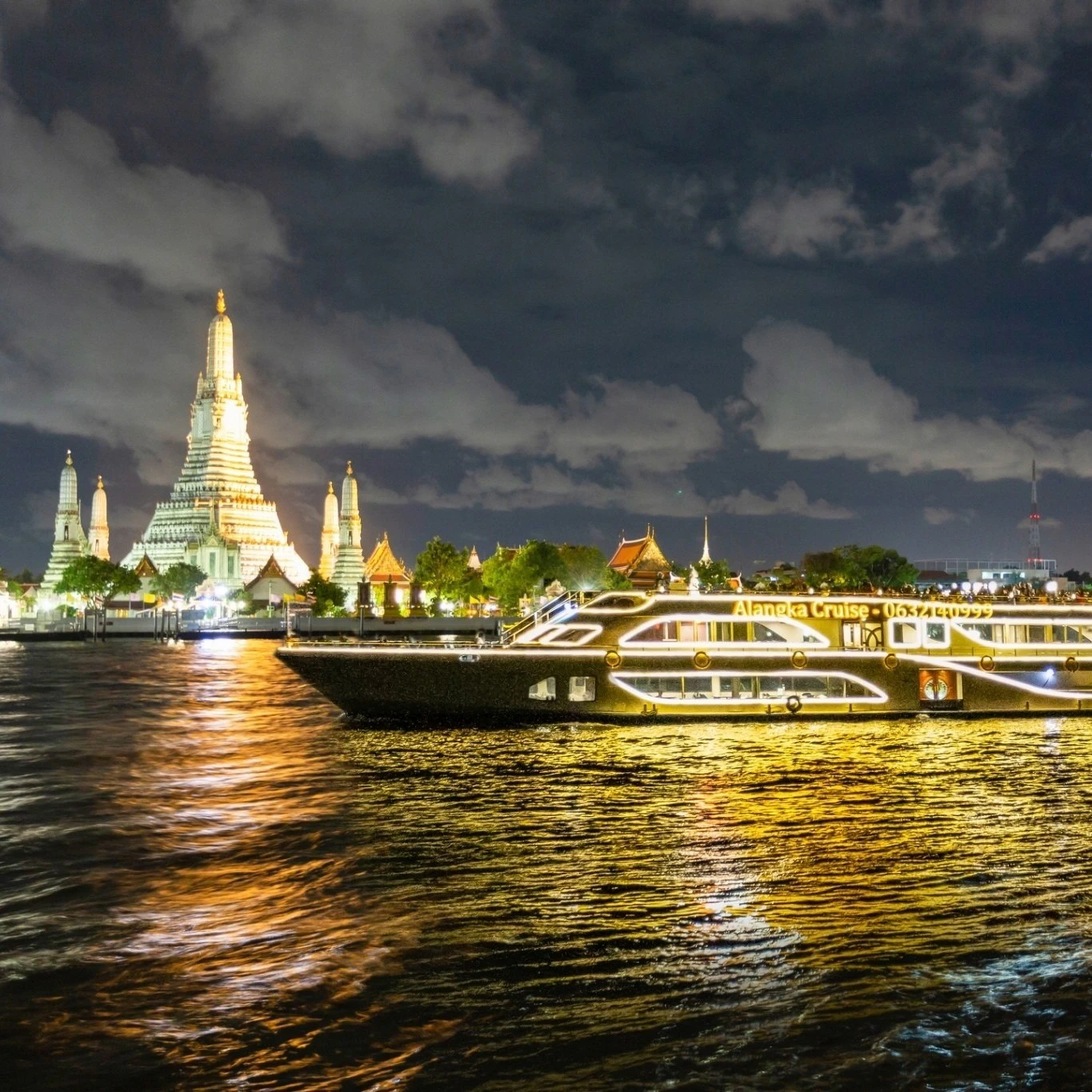 A brightly lit cruise ship on a river at night with a large illuminated temple and other buildings in the background.