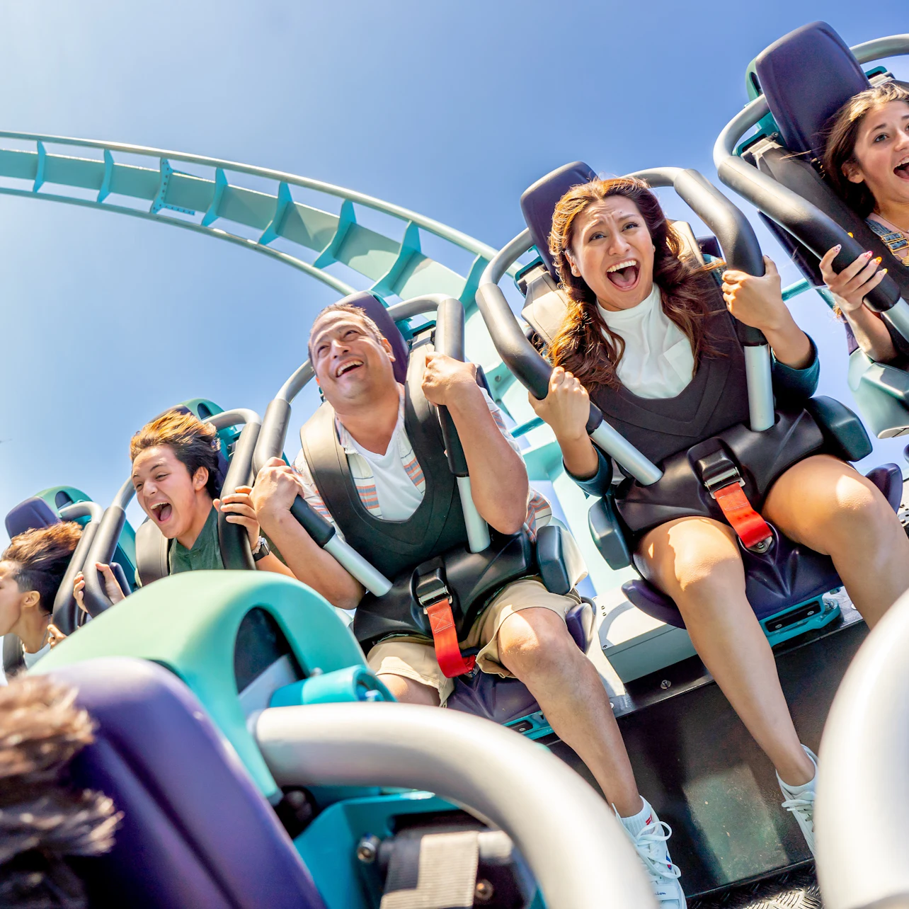 Four people screaming with excitement on a rollercoaster with a blue track against a clear sky.