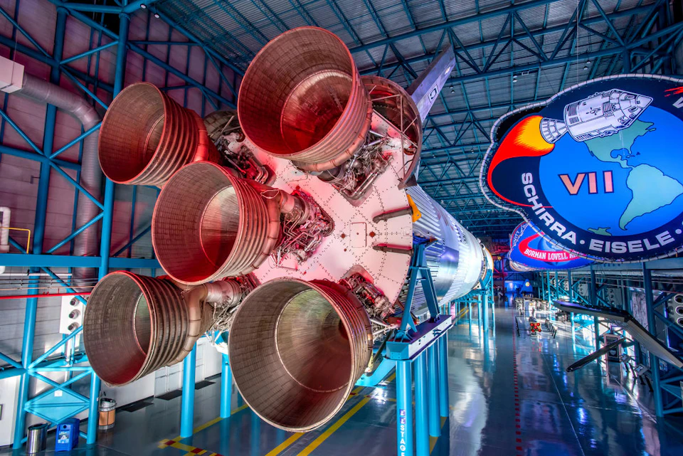The image shows the large engines of a Saturn V rocket displayed indoors with signage and metallic structures surrounding it.