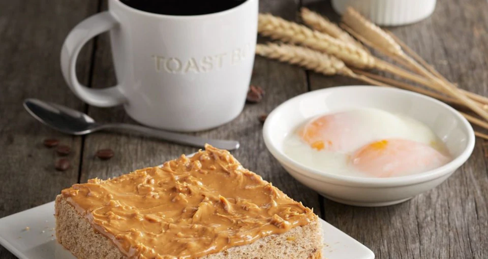 A slice of bread with peanut butter, a bowl with two boiled eggs, wheat stalks, and a white mug with "TOAST" embossed on it.