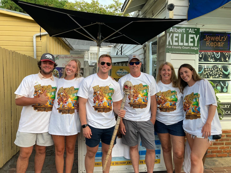 A group of five people wearing matching themed T-shirts pose together outdoors near a wooden fence and a building.