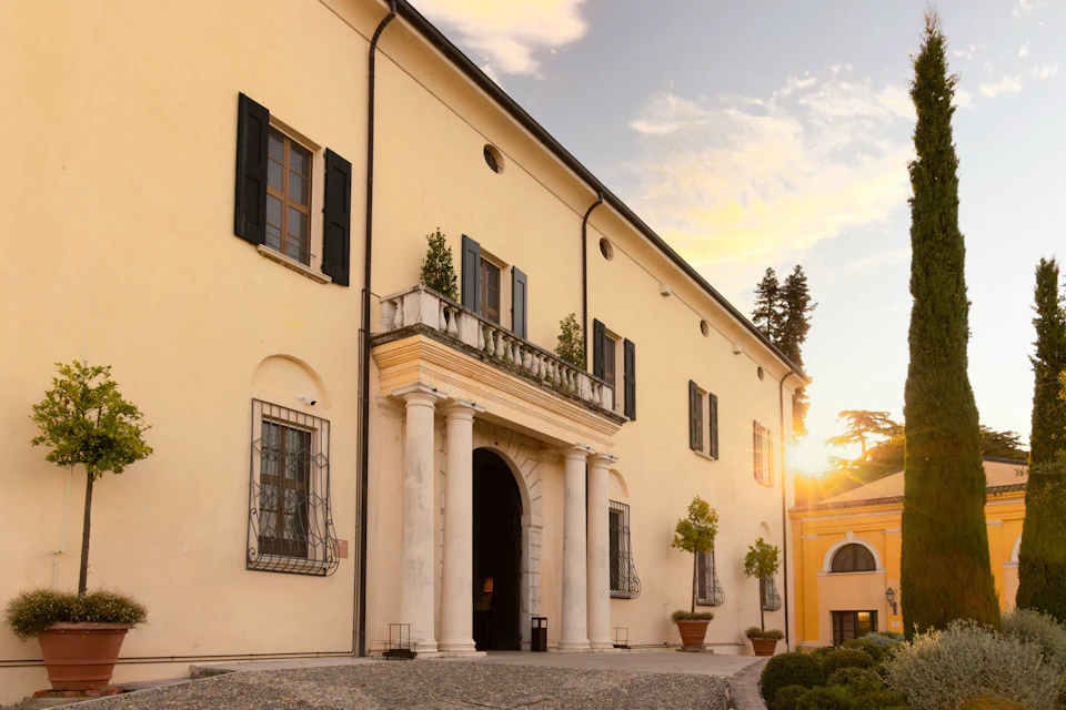 A beige building with classical columns at the entrance, black shutters, and potted plants, under a sunset sky.