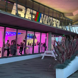 Människor samlas i en neonbelyst lokal med glasväggar och modern inredning. Exteriören har stora planteringslådor och en vit bänk.