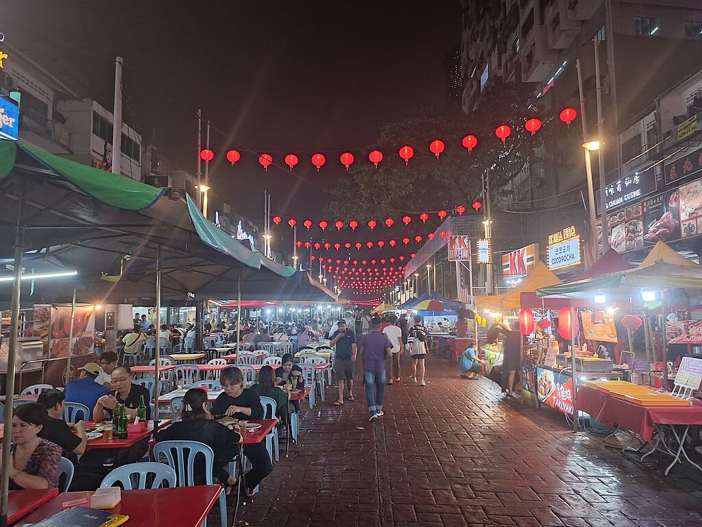 Night market with food stalls, red lanterns hanging above, and people dining at tables along a busy street.