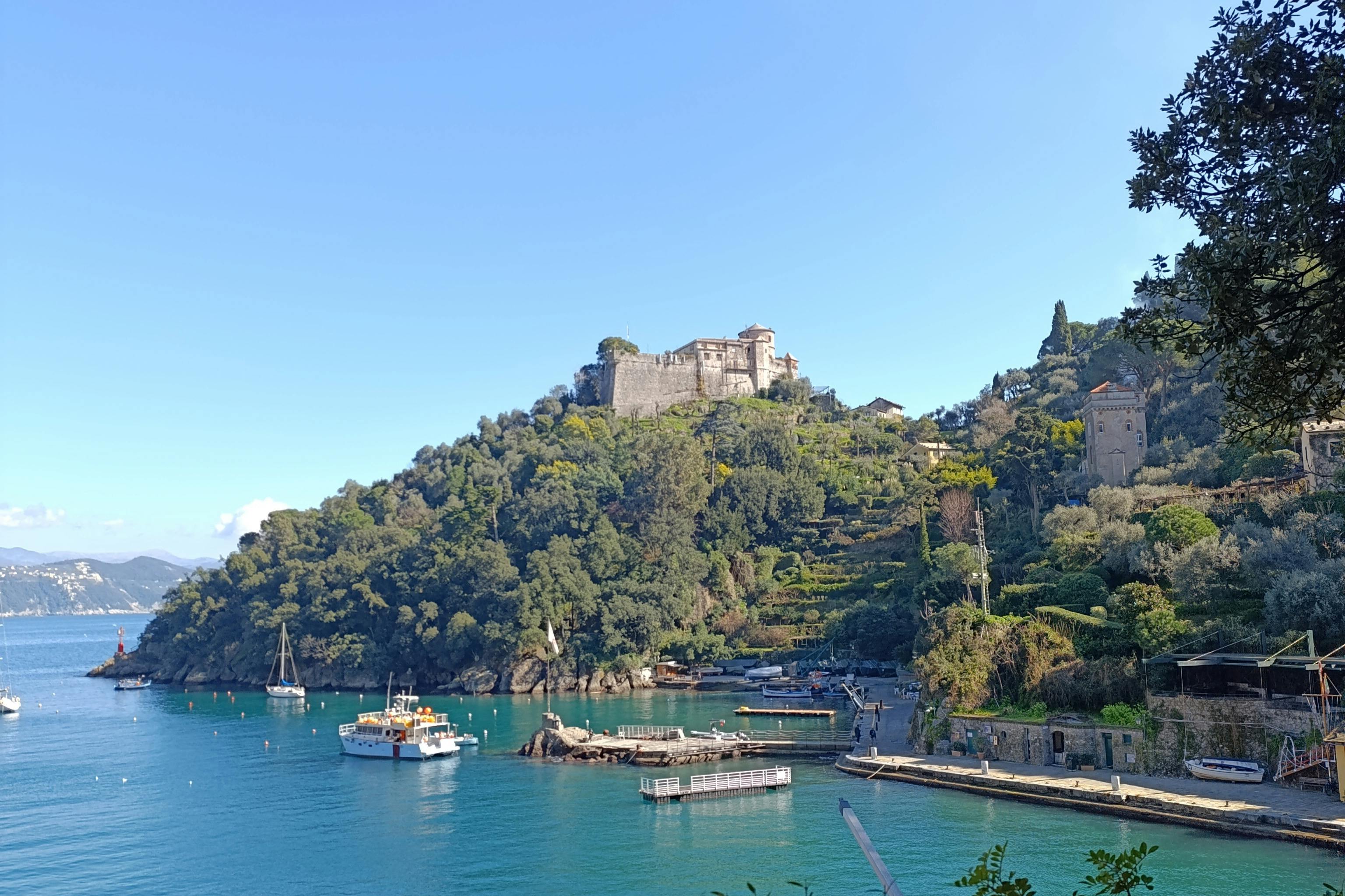 A coastal hill with a castle at the top, boats docked in turquoise water below, and lush greenery on the slopes.