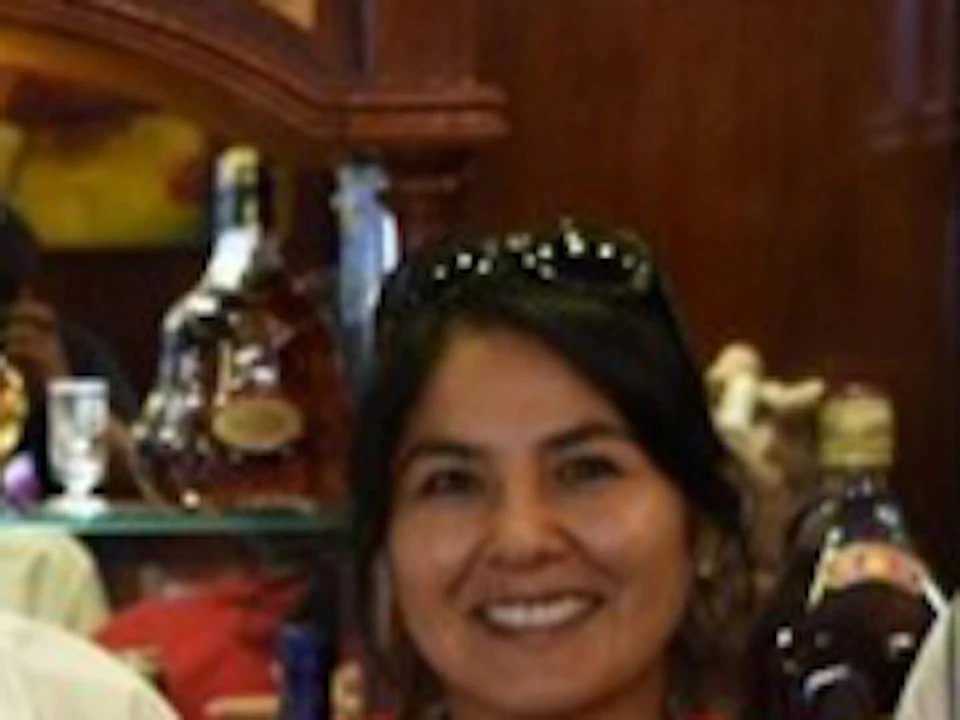 Three people stand behind a bar, smiling. The bar counter displays several drinks and a tray of eggs. Shelves stocked with bottles are behind them.