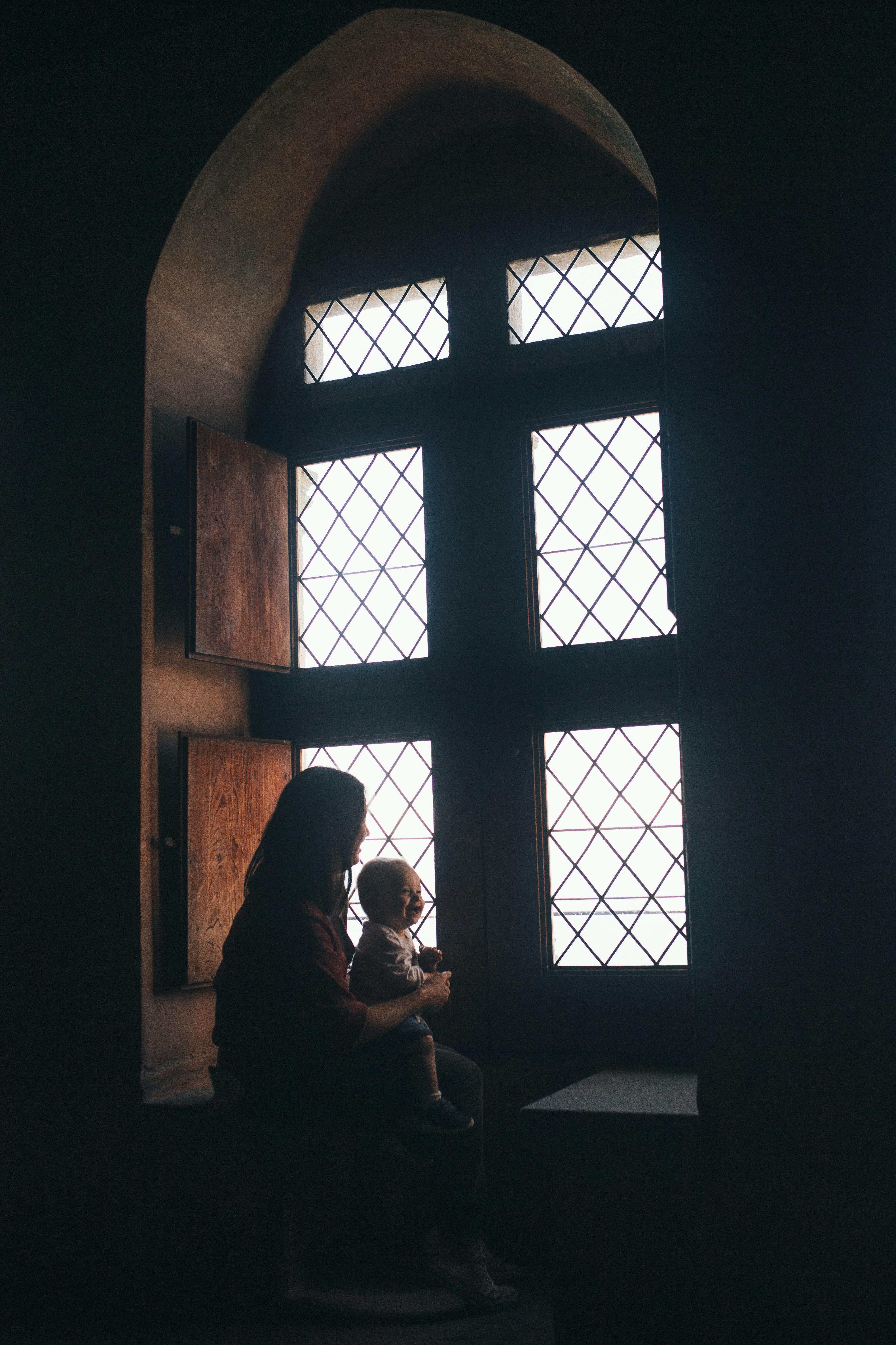 Silhouetted adult and baby sit by a large, grid-paned window with wooden shutters in a dimly lit room.