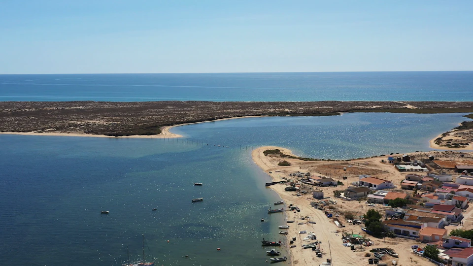 Aerial view of a coastal landscape with a sandy beach, several boats anchored in the water, and buildings near the shoreline.