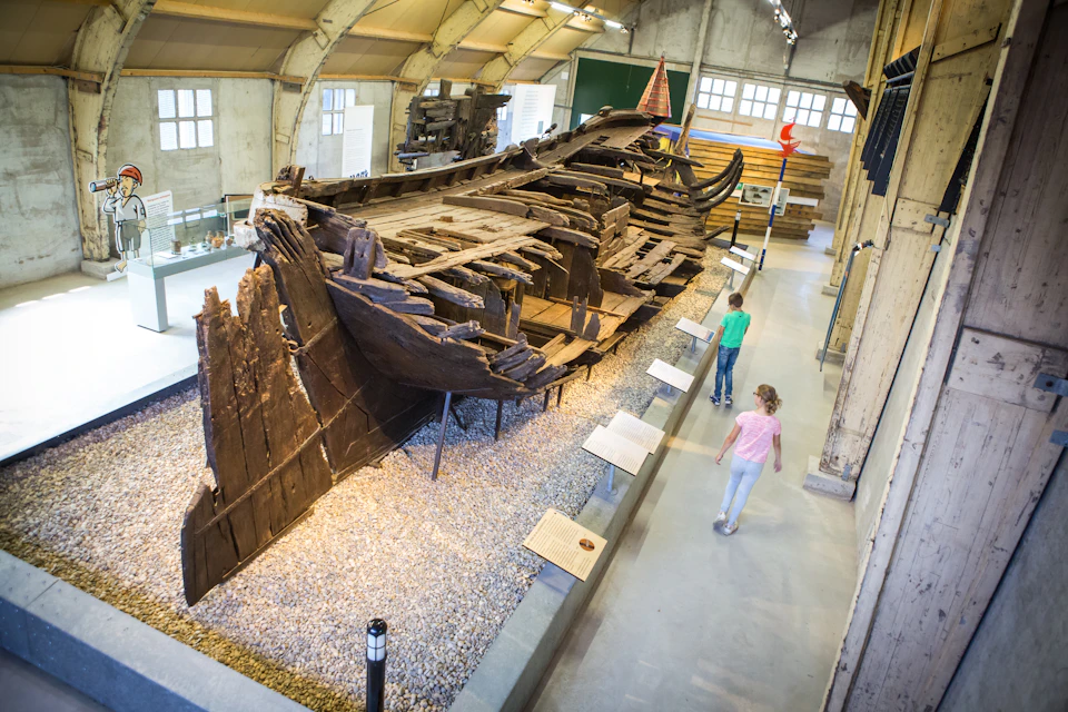 Two children walk past a large, ancient wooden shipwreck displayed indoors on a gravel bed with information plaques nearby.