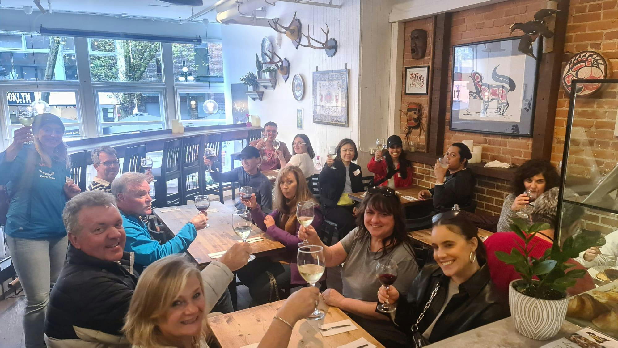 Grupa osób siedzących przy drewnianym stole w rustykalnej restauracji, podnoszących kieliszki wina i uśmiechających się do kamery.