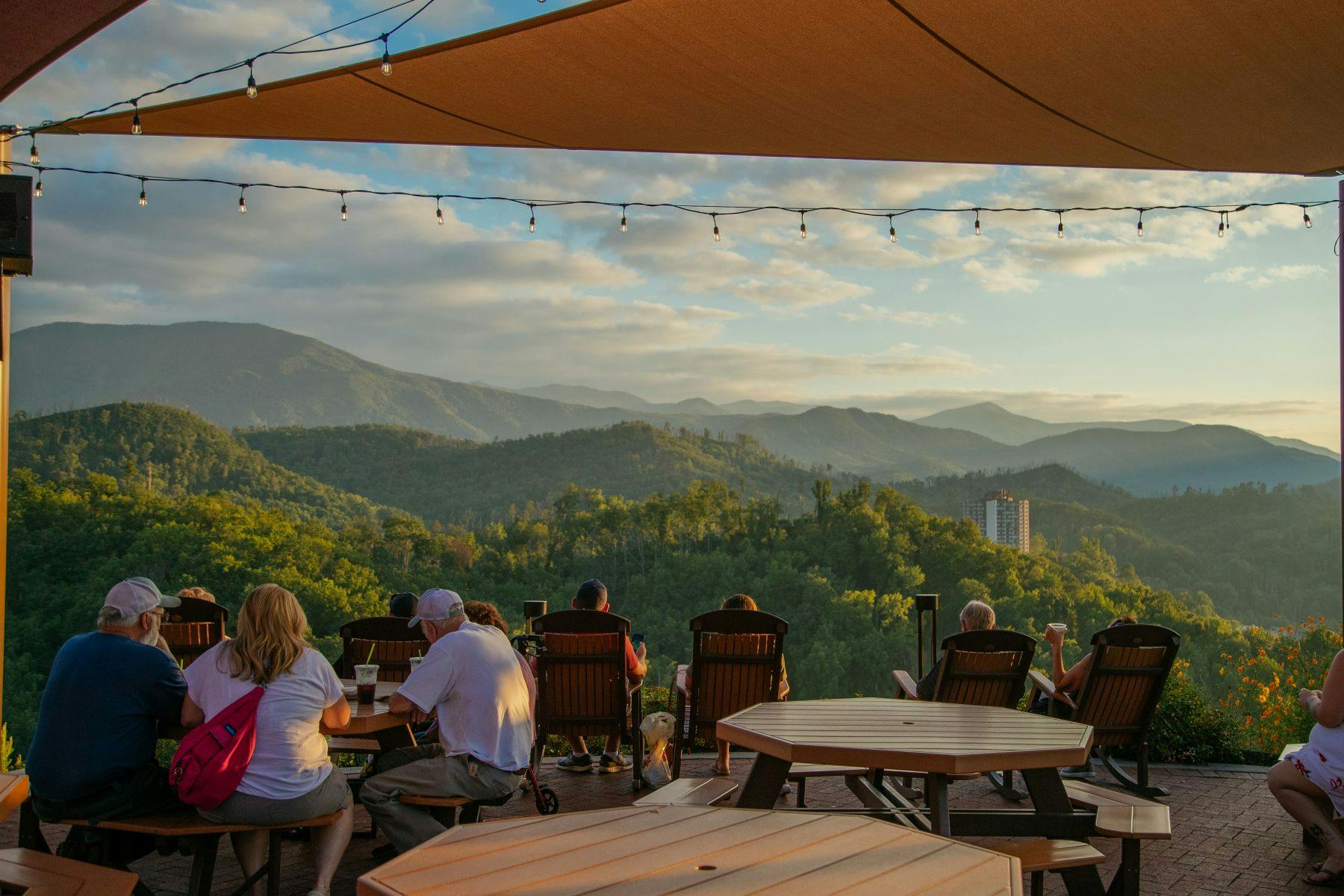 People seated on a deck with wooden tables and chairs, enjoying a mountain view under a canopy with string lights.