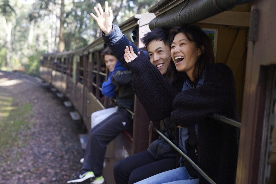 Deux personnes souriant et faisant des signes de la main, assises sur le côté d'un wagon de train ouvert, avec un arrière-plan boisé visible.