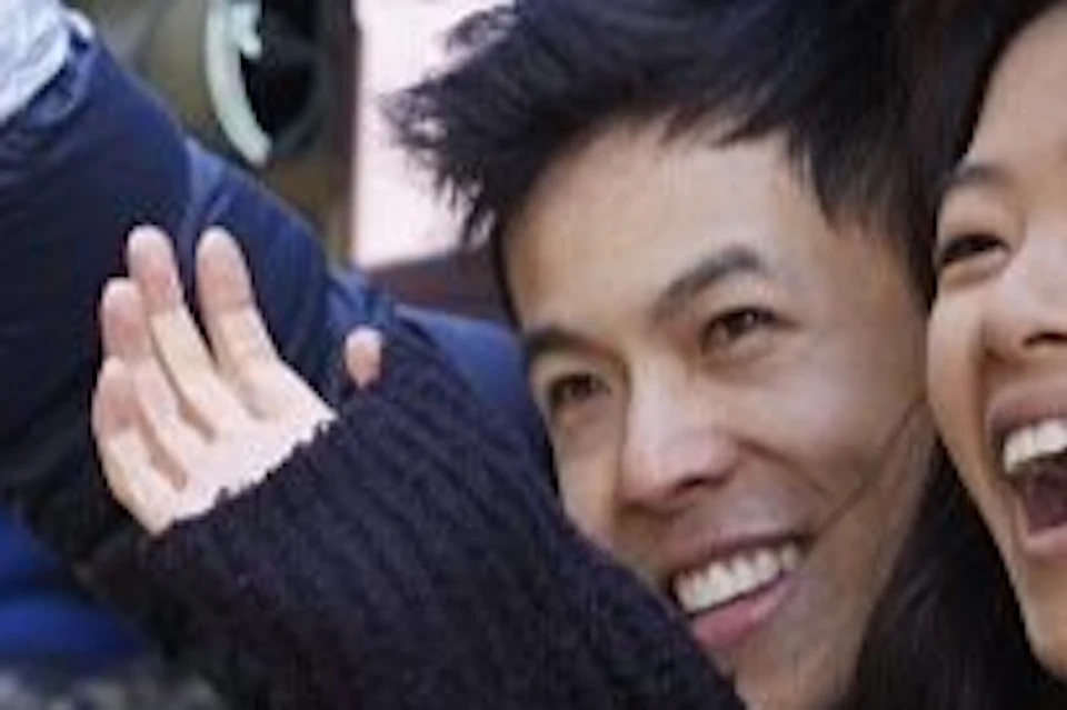 Two people smiling and waving while sitting on the side of an open train carriage, with a wooded background visible.