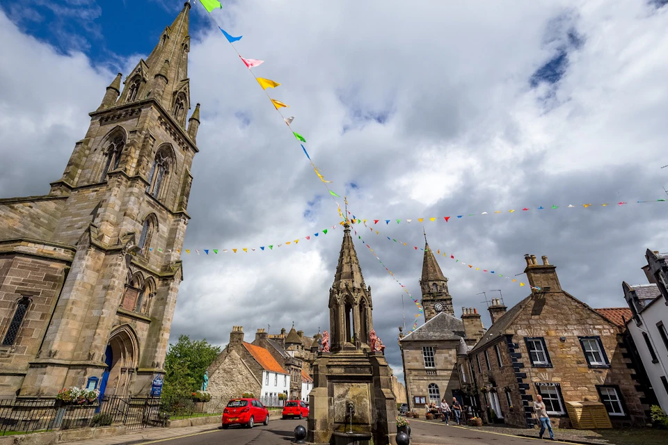Town square with a stone monument, colorful bunting, historic buildings, and a church steeple under a cloudy sky.